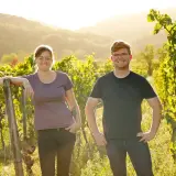 Zwei junge Winzer aus der Pfalz lächeln in der goldenen Abendsonne inmitten ihrer Weinreben.