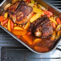 Zwei saftige Stücke **Schweinebraten mit Rotwein** schmoren in einer Bratpfanne mit Kartoffeln und Karotten im Ofen.