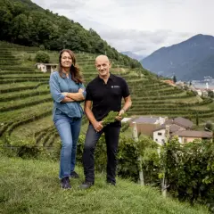 Ein Paar steht vor terrassierten Weinbergen am Lago di Lugano, mit Blick auf das Dorf Morcote. Das Castello di Morcote ist in der Ferne zu erahnen.