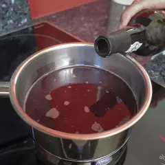 Eine Hand gießt Rotwein in einen Topf, um ihn zu kochen. Der Herd zeigt die Einstellung "LO".