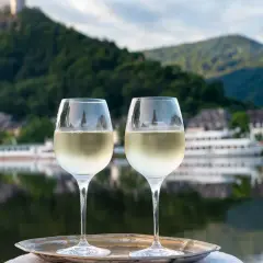 Zwei Gläser Weißwein auf einem Tablett vor einem malerischen Weinbaugebiet am Rhein mit Burg und Fluss.