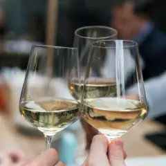 Drei Gläser mit hellem Wein, ein Sauvignon Blanc trocken, werden zum Anstoßen gehalten. Im Hintergrund verschwimmen die Silhouetten von Menschen.
