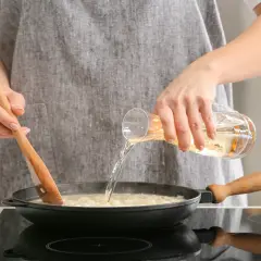 Eine Person verkocht Alkohol beim Kochen. Sie gießt eine Flüssigkeit aus einem Glas in eine Pfanne, während sie mit einem Holzlöffel umrührt.