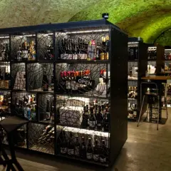 Weinregale mit Flaschen und Stehtischen in einem Gewölbekeller mit grüner Beleuchtung.