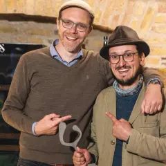 Zwei Männer mit Hüten und Brillen halten eine graue Skulptur. Sie zeigen darauf und lächeln. Ein Zeichen für "Wein gegen Rassismus".