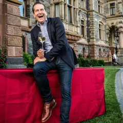 Gerhard Retter lacht ausgelassen mit einem Glas Wein in der Hand vor einem Schloss.