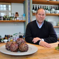 Otto Geisel sitzt an einem Holztisch mit Artischocken. Regale voller Gläser und Flaschen sind im Hintergrund zu sehen.