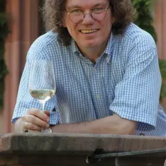 Ernst Loosen, mit lockigem Haar und Brille, hält ein Glas Weißwein. Er trägt ein blau-weiß kariertes Hemd und lächelt.