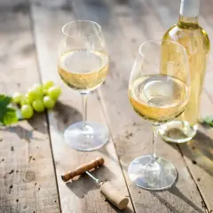 Zwei Gläser Weißwein, eine Flasche, Trauben und ein Korkenzieher auf Holztisch. Der Unterschied Riesling Chardonnay wird hier visuell angedeutet.