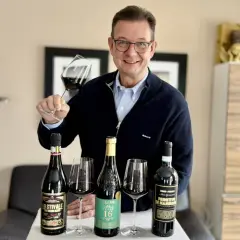 Luca Maroni Punkte: Mann mit Brille hält Weinglas, daneben drei Flaschen Rotwein und Gläser.