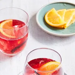 Zwei erfrischende Cocktails mit Portwein, garniert mit Orangenscheiben und Zimtstangen, stehen bereit. Daneben ein Teller mit frischen Orangenspalten.