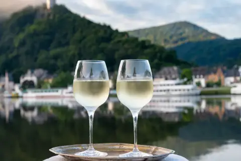 Zwei Gläser Weißwein auf einem Tablett vor einem malerischen Weinbaugebiet am Rhein mit Burg und Fluss.