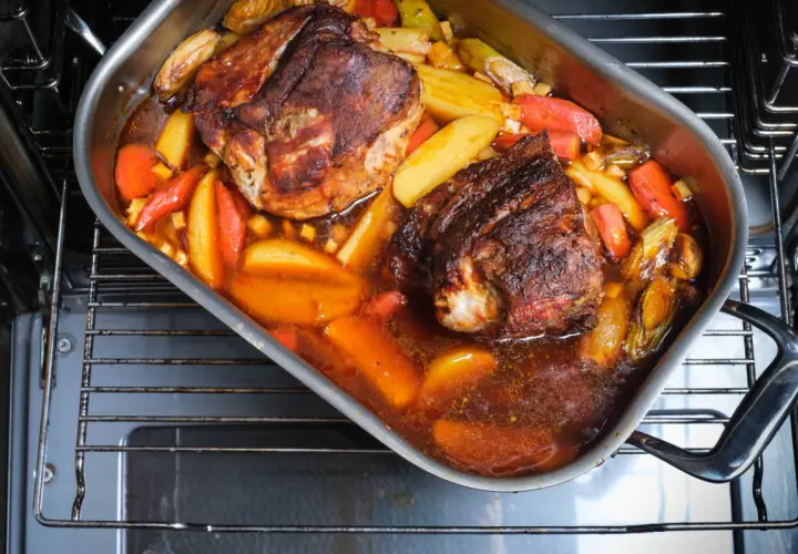 Zwei saftige Stücke **Schweinebraten mit Rotwein** schmoren in einer Bratpfanne mit Kartoffeln und Karotten im Ofen.