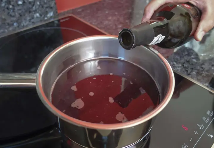 Eine Hand gießt Rotwein in einen Topf, um ihn zu kochen. Der Herd zeigt die Einstellung "LO".