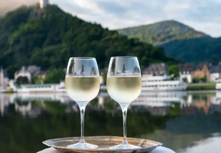 Zwei Gläser Weißwein auf einem Tablett vor einem malerischen Weinbaugebiet am Rhein mit Burg und Fluss.