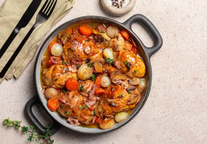 Herzhaftes Coq au Vin in einer Pfanne, mit Hähnchenteilen, Karotten, Zwiebeln und Pilzen in einer reichhaltigen Sauce.