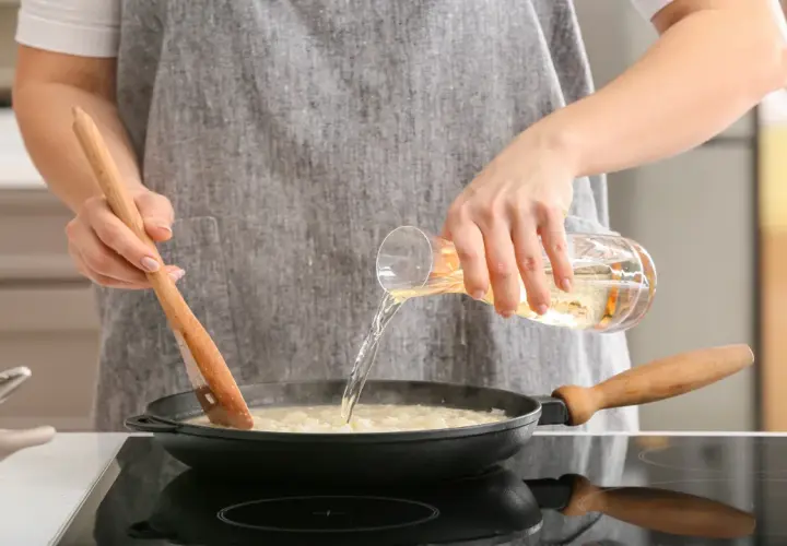 Eine Person verkocht Alkohol beim Kochen. Sie gießt eine Flüssigkeit aus einem Glas in eine Pfanne, während sie mit einem Holzlöffel umrührt.