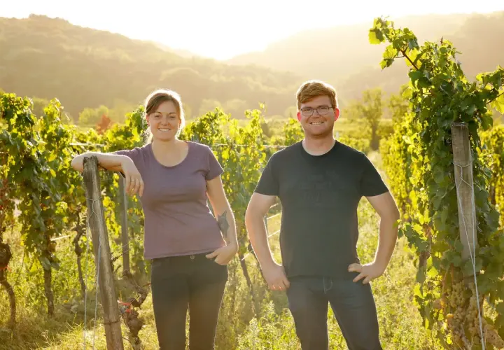 Zwei junge Winzer aus der Pfalz lächeln in der goldenen Abendsonne inmitten ihrer Weinreben.