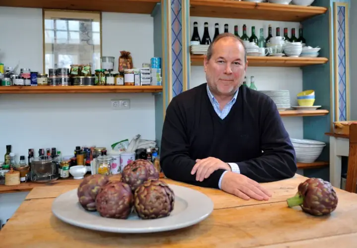 Otto Geisel sitzt an einem Holztisch mit Artischocken. Regale voller Gläser und Flaschen sind im Hintergrund zu sehen.