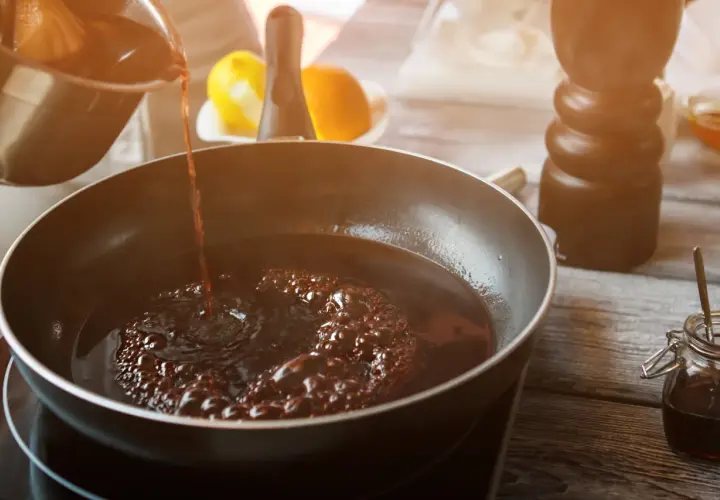Dunkler Portwein zum Kochen wird in eine Pfanne gegossen, wo er zu köcheln beginnt. Zitrusfrüchte und ein Gewürzstreuer sind im Hintergrund zu sehen.