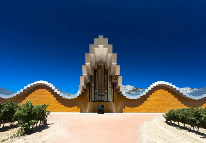 Moderne Weinbodega mit wellenförmiger Fassade und metallischem Dach in Rioja Alavesa, umgeben von Weinbergen und Bergen.