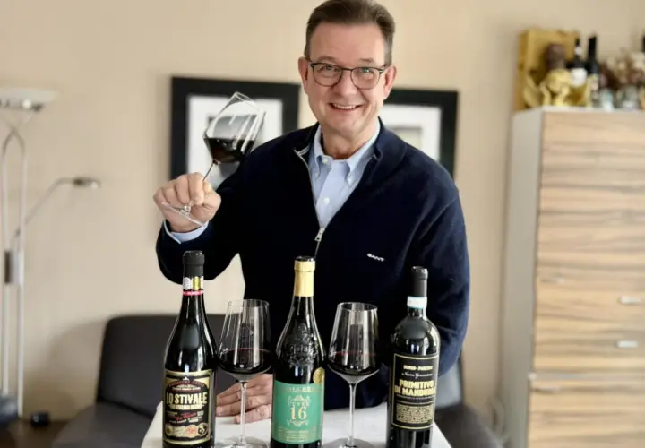 Luca Maroni Punkte: Mann mit Brille hält Weinglas, daneben drei Flaschen Rotwein und Gläser.