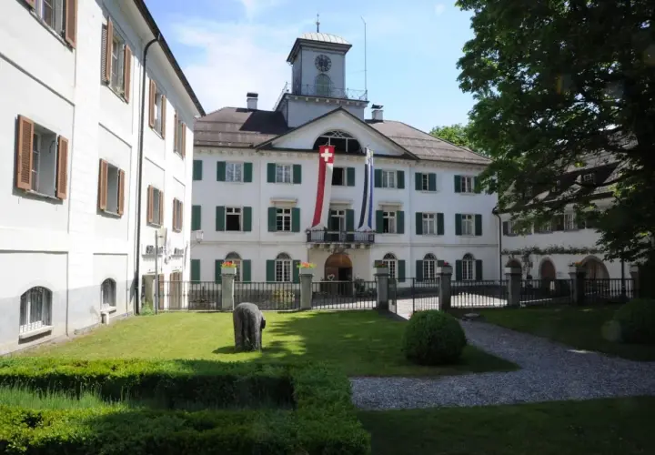 Das Schloss Reichenau mit Schweizer Fahnen und einem Uhrturm. Ein gepflegter Garten mit einem Stein und Büschen lädt zum Verweilen ein.