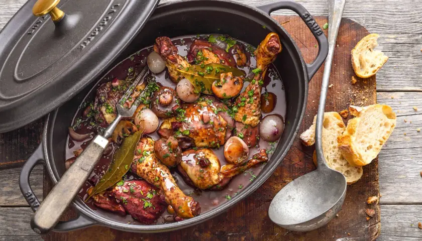 Ein köstliches Coq au Vin ganzes Huhn in dunkler Sauce mit Pilzen und Zwiebeln, serviert in einem Gusseisentopf. Dazu Baguette.