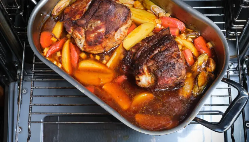 Zwei saftige Stücke **Schweinebraten mit Rotwein** schmoren in einer Bratpfanne mit Kartoffeln und Karotten im Ofen.