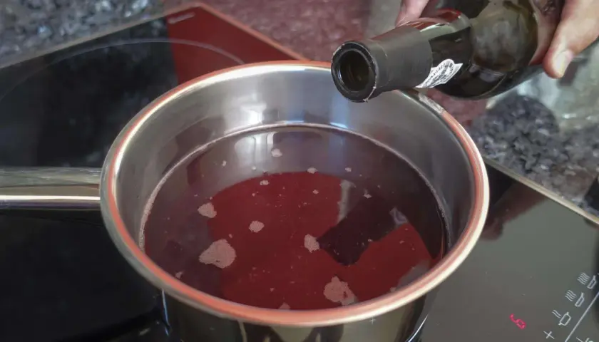 Eine Hand gießt Rotwein in einen Topf, um ihn zu kochen. Der Herd zeigt die Einstellung "LO".