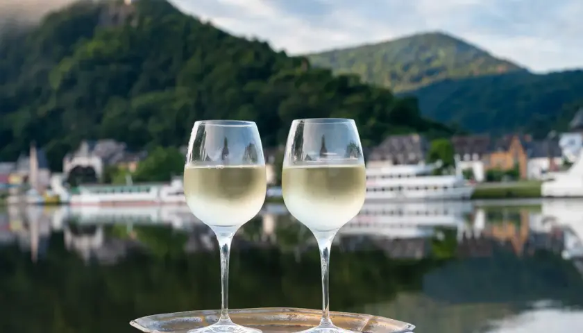 Zwei Gläser Weißwein auf einem Tablett vor einem malerischen Weinbaugebiet am Rhein mit Burg und Fluss.