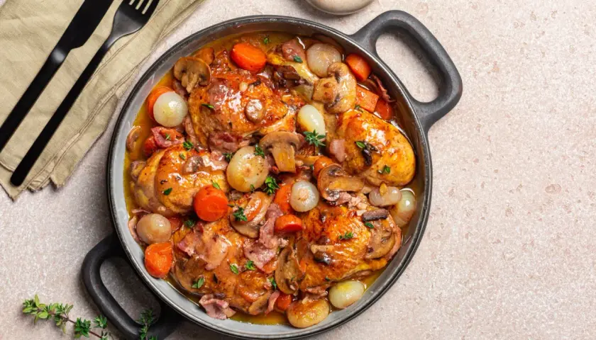 Herzhaftes Coq au Vin in einer Pfanne, mit Hähnchenteilen, Karotten, Zwiebeln und Pilzen in einer reichhaltigen Sauce.