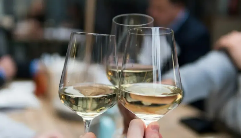Drei Gläser mit hellem Wein, ein Sauvignon Blanc trocken, werden zum Anstoßen gehalten. Im Hintergrund verschwimmen die Silhouetten von Menschen.