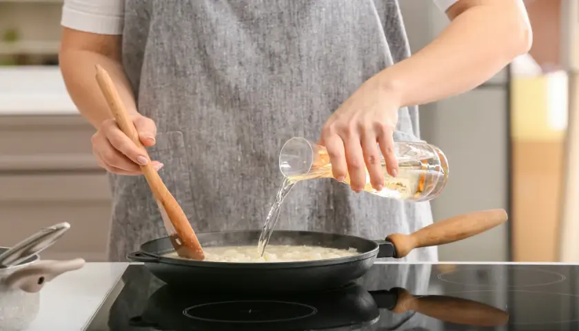 Eine Person verkocht Alkohol beim Kochen. Sie gießt eine Flüssigkeit aus einem Glas in eine Pfanne, während sie mit einem Holzlöffel umrührt.