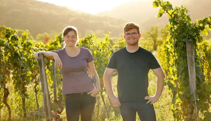 Zwei junge Winzer aus der Pfalz lächeln in der goldenen Abendsonne inmitten ihrer Weinreben.
