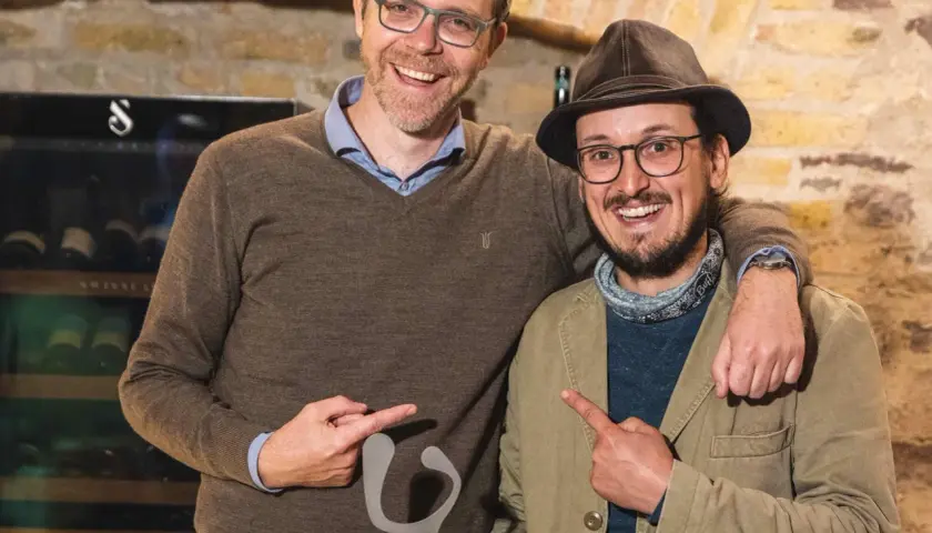 Zwei Männer mit Hüten und Brillen halten eine graue Skulptur. Sie zeigen darauf und lächeln. Ein Zeichen für "Wein gegen Rassismus".