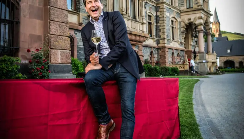 Gerhard Retter lacht ausgelassen mit einem Glas Wein in der Hand vor einem Schloss.