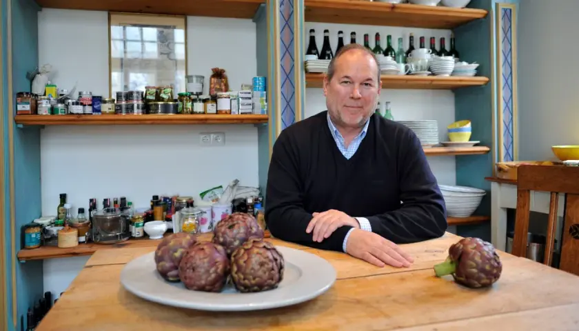 Otto Geisel sitzt an einem Holztisch mit Artischocken. Regale voller Gläser und Flaschen sind im Hintergrund zu sehen.