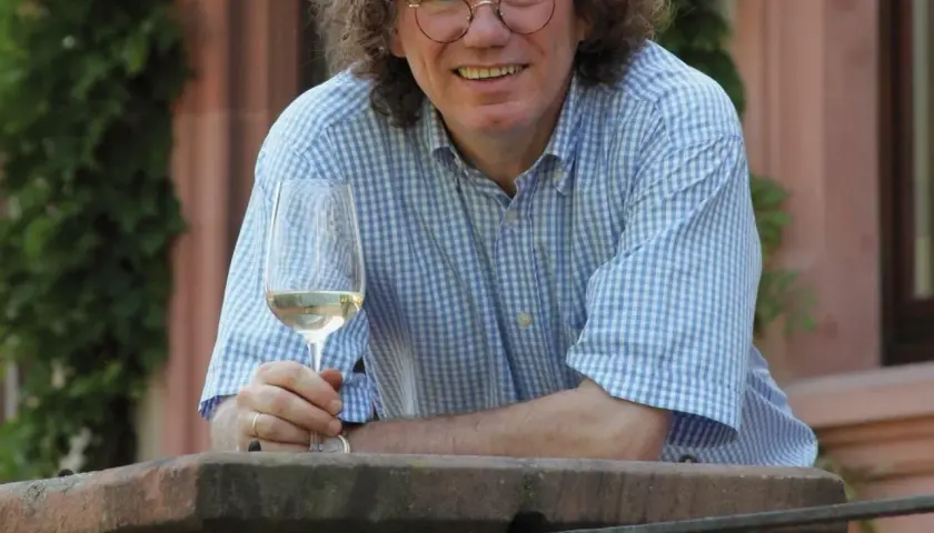 Ernst Loosen, mit lockigem Haar und Brille, hält ein Glas Weißwein. Er trägt ein blau-weiß kariertes Hemd und lächelt.