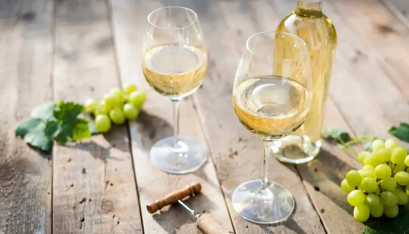 Zwei Gläser Weißwein, eine Flasche, Trauben und ein Korkenzieher auf Holztisch. Der Unterschied Riesling Chardonnay wird hier visuell angedeutet.