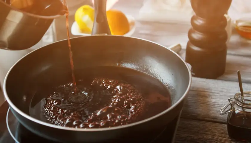 Dunkler Portwein zum Kochen wird in eine Pfanne gegossen, wo er zu köcheln beginnt. Zitrusfrüchte und ein Gewürzstreuer sind im Hintergrund zu sehen.