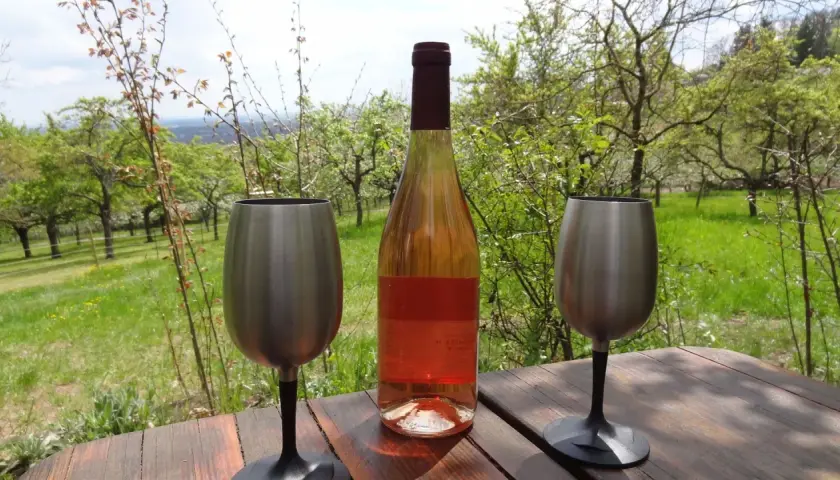 Flasche Roséwein und zwei Edelstahlgläser auf Holztisch, umgeben von blühenden Obstbäumen. Ein echter Geheimtipp von einem Winzer aus der Pfalz.