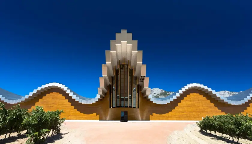 Moderne Weinbodega mit wellenförmiger Fassade und metallischem Dach in Rioja Alavesa, umgeben von Weinbergen und Bergen.