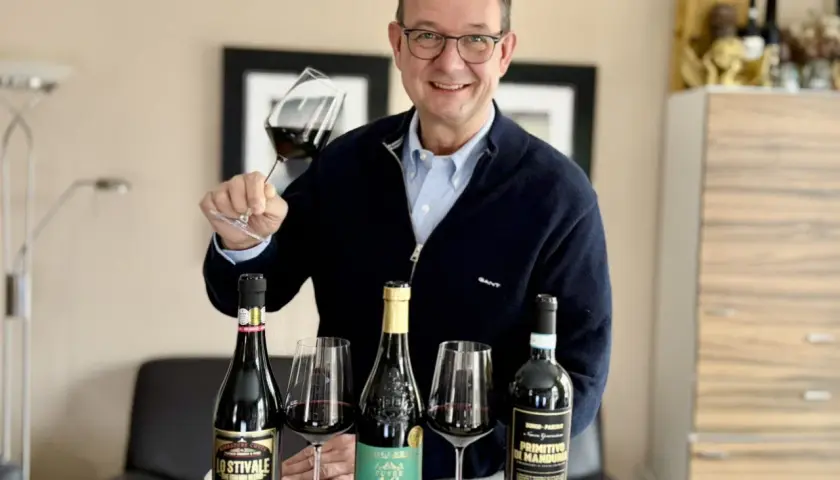 Luca Maroni Punkte: Mann mit Brille hält Weinglas, daneben drei Flaschen Rotwein und Gläser.