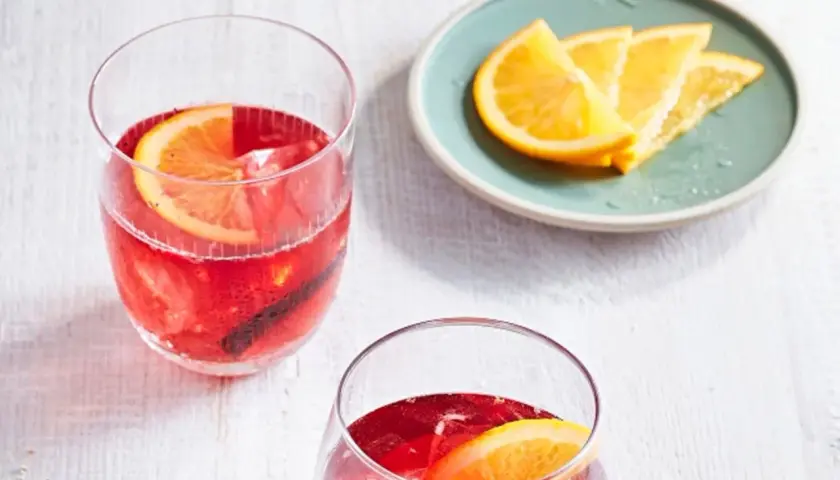 Zwei erfrischende Cocktails mit Portwein, garniert mit Orangenscheiben und Zimtstangen, stehen bereit. Daneben ein Teller mit frischen Orangenspalten.