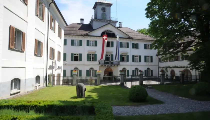 Das Schloss Reichenau mit Schweizer Fahnen und einem Uhrturm. Ein gepflegter Garten mit einem Stein und Büschen lädt zum Verweilen ein.