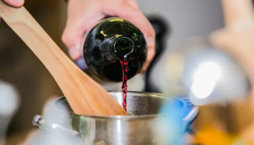 Rotwein wird in einen Topf gegossen, während ein Kochlöffel daneben liegt. Die Frage, wie lange braucht Rotwein zum Verkochen, ist hier relevant.