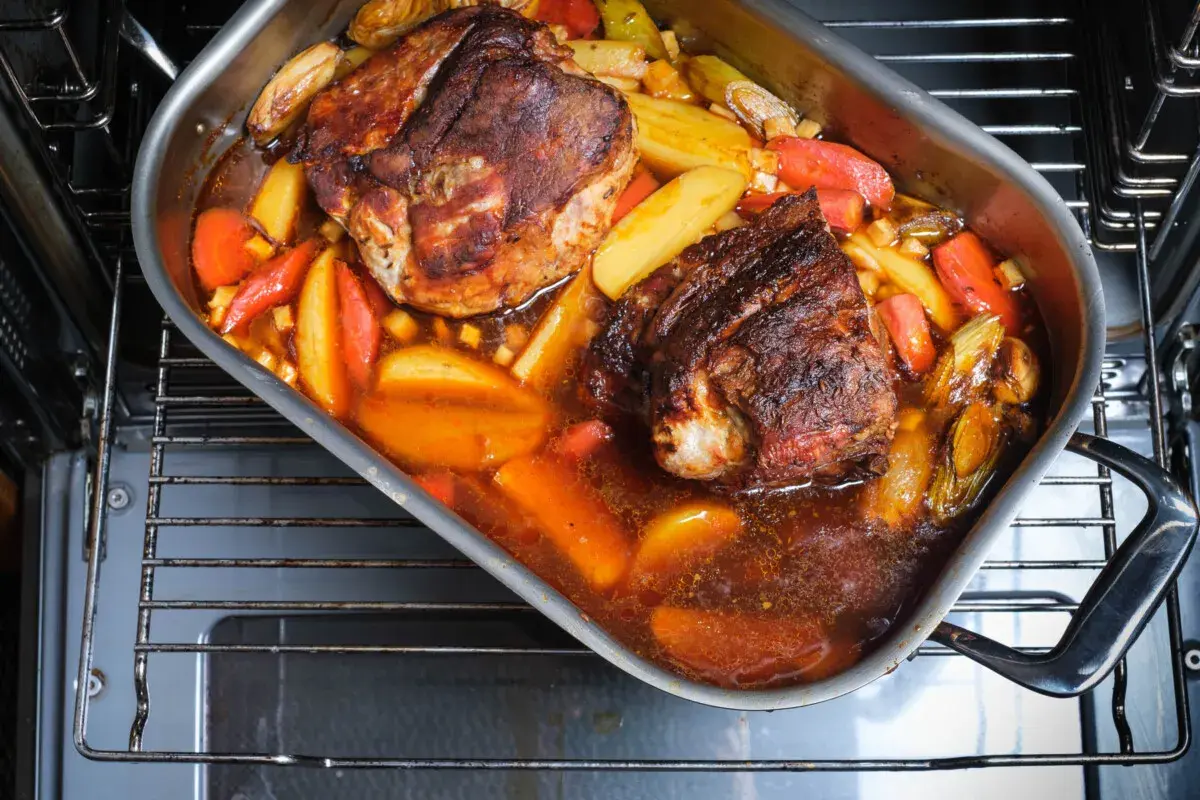 Zwei saftige Stücke **Schweinebraten mit Rotwein** schmoren in einer Bratpfanne mit Kartoffeln und Karotten im Ofen.