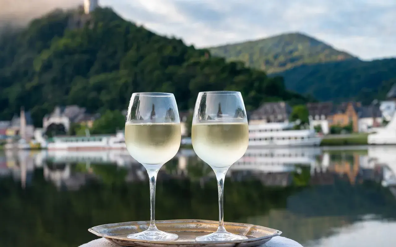 Zwei Gläser Weißwein auf einem Tablett vor einem malerischen Weinbaugebiet am Rhein mit Burg und Fluss.