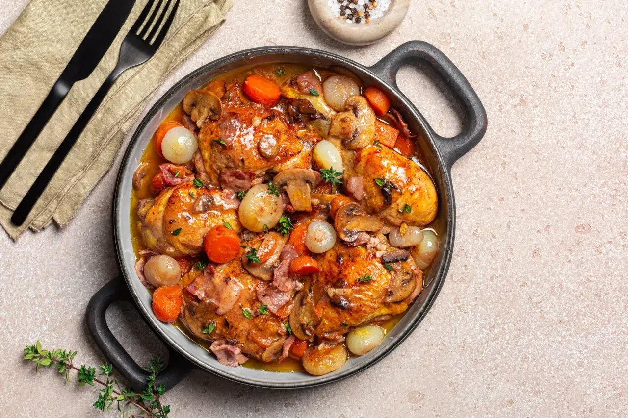 Herzhaftes Coq au Vin in einer Pfanne, mit Hähnchenteilen, Karotten, Zwiebeln und Pilzen in einer reichhaltigen Sauce.